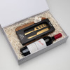 A wine bottle, a wooden cheese board, and a gold-handled cheese knife in a gift box with shredded paper.