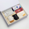 A gift box containing a striped cloth, two coffee mugs (one white with a cork base, one black), and shortbread biscuits.