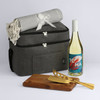 A cooler bag in grey with a blanket, a bottle of white wine, a wooden cutting board, and gold utensils.
