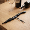 A black wine waiters knife positioned on a wooden table, alongside a wine bottle and a glass.