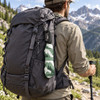 A backpack with a green and white patterned drink bottle attached via a carabiner, set against a mountainous landscape.