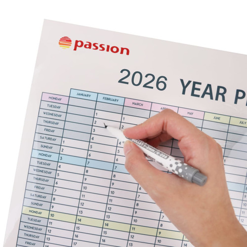 A hand holding a grey pen marks a date on a colourful 2026 year planner with a logo in the corner.