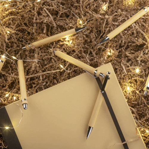 A collection of bamboo pens in natural colour scattered around a notebook and decorative lights on brown filler material.