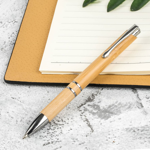 A bamboo pen with metal accents resting on a textured notebook, surrounded by greenery.