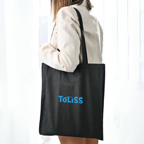 A black shopping tote bag with a V gusset, featuring a logo, held by a person against a light backdrop.