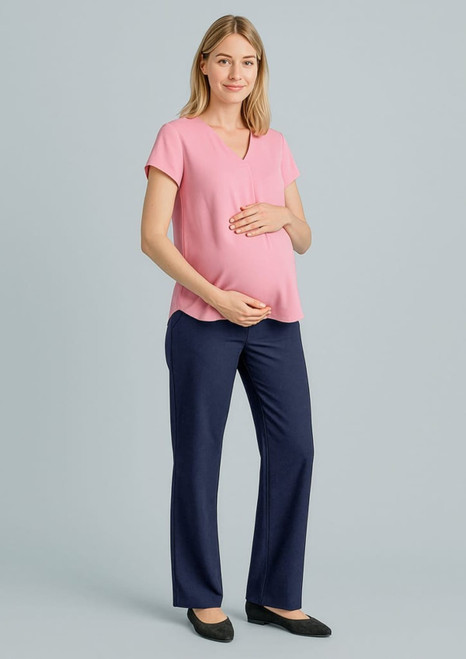 A pregnant woman wears a pink top and navy maternity pants, standing with one hand on her belly. The background is neutral.