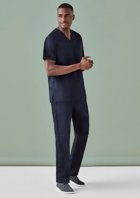 A man is wearing navy unisex scrub pants and a matching scrub top, standing against a plain green background.
