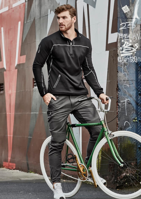 A man in black pullover and grey cuffed stretch pants poses with a green bicycle against a graffiti wall.