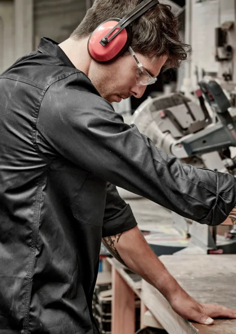 A man wearing a black long sleeve shirt, safety glasses, and earmuffs, working at a bench with machinery.