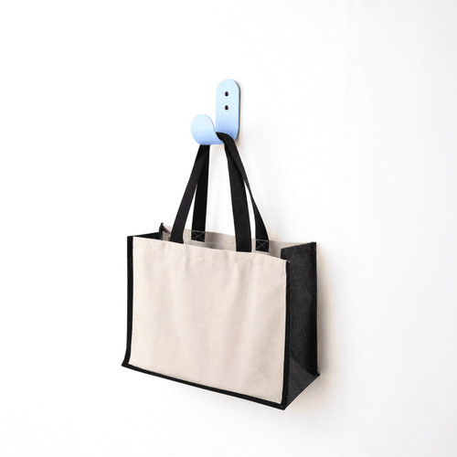 A functional tote bag in beige and black hangs on a blue hook against a white wall. The bag has handles and a logo.