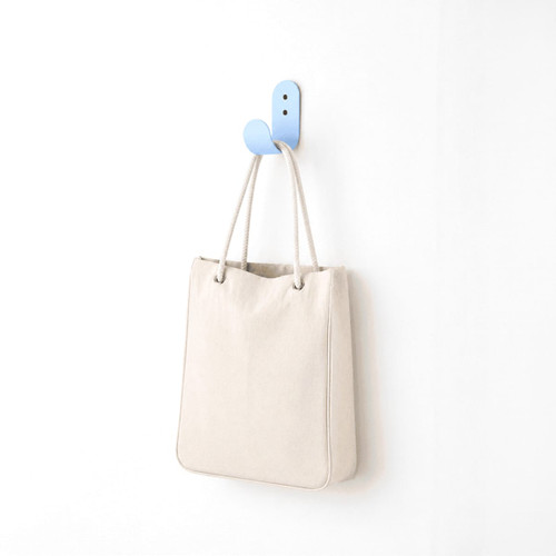 A cream-coloured tote bag hanging on a blue hook against a white wall. The bag has a simple design and a logo.
