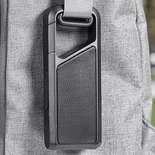 A portable black Bluetooth speaker attached to a grey bag with a strap. It has a textured surface and a prominent speaker grille.