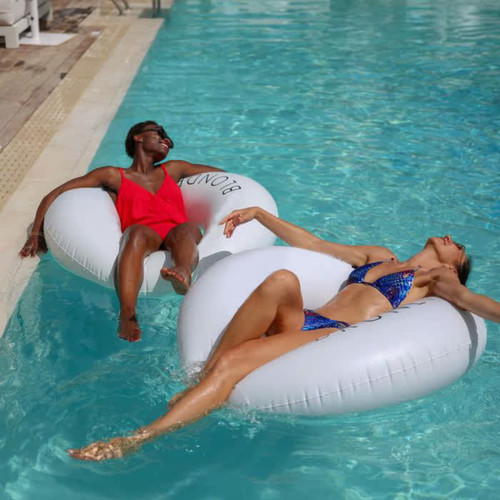 Two women relax in white inflatable swim rings in a pool, surrounded by blue water and sunlight.