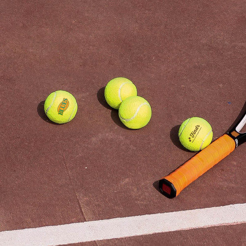 Three bright yellow tennis balls and a tennis racquet resting on a brown court surface.