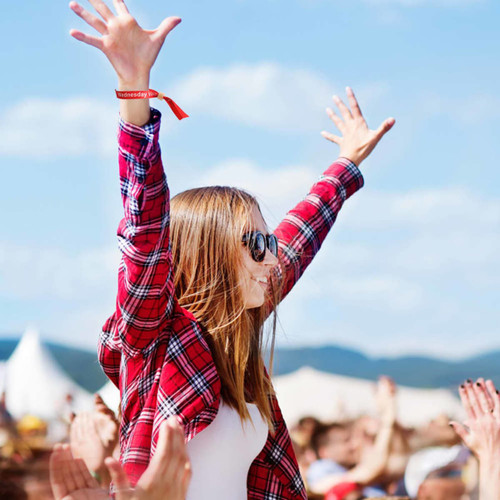 A woman with long hair in sunglasses and a red plaid shirt raises her arms, wearing a vibrant wristband at an outdoor event.
