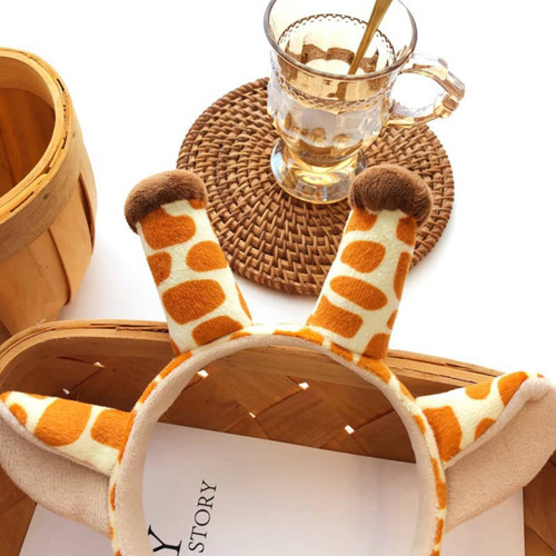 A giraffe-themed ear hairband in orange and white with plush ears, displayed alongside a glass and woven mat.