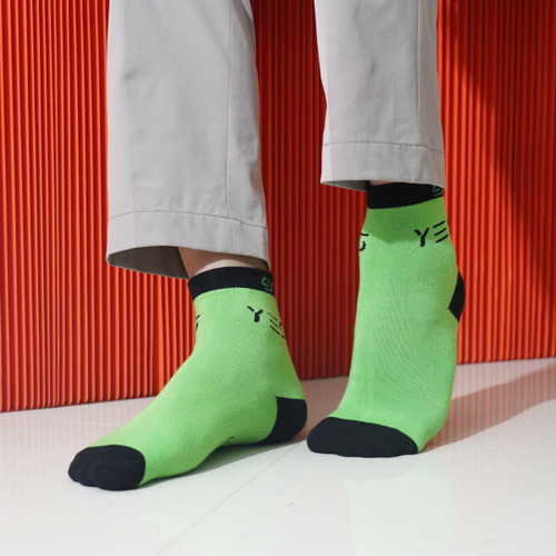 Bright green ankle socks with black accents and a logo, positioned on feet against a vibrant orange backdrop.