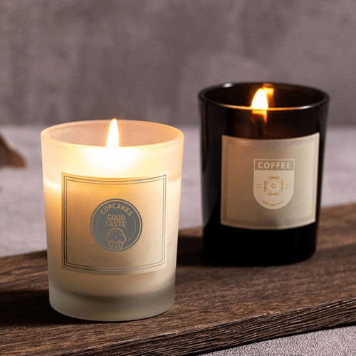 A frosted white candle and a black candle, both lit, sitting on a wooden surface with labels.