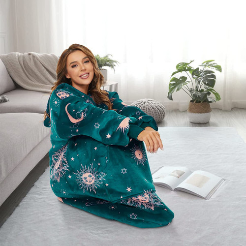 A woman sits on a rug wearing a teal wearable blanket featuring a celestial design of stars and sun motifs.