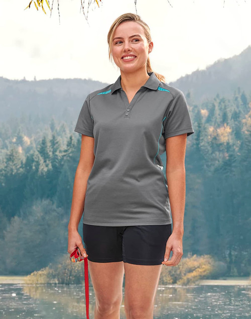 A woman stands outdoors wearing a grey polo shirt with blue contrast accents, alongside black shorts and holding a leash.