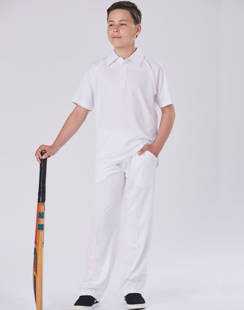 A boy stands confidently wearing white cricket pants and a matching polo shirt, holding a cricket bat.
