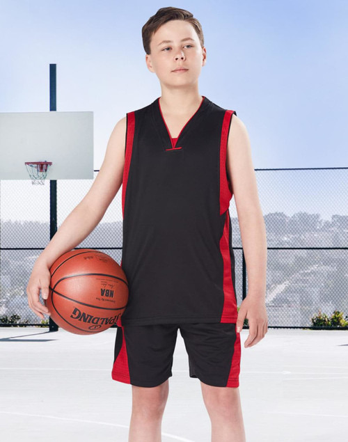 A young boy stands on a basketball court wearing black and red basketball shorts and a matching sleeveless top, holding a basketball.
