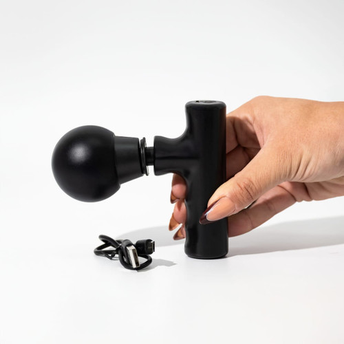 A black mini massage gun being held by a hand, alongside a charging cable.