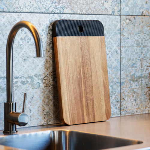 A wooden serving board with a black handled edge, leaning against a tiled wall near a sink.