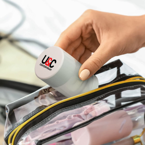 A grey travel bottle being placed into a transparent zippered bag, with other items visible inside.