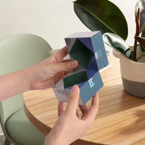 A person holds a 3D Flips Diamond Magic Cube in shades of blue and green, placed on a wooden table beside a plant.