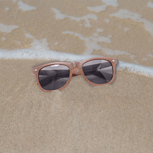 Coffee fibre sunglasses in a brown hue resting on sandy beach, with gentle waves in the background.