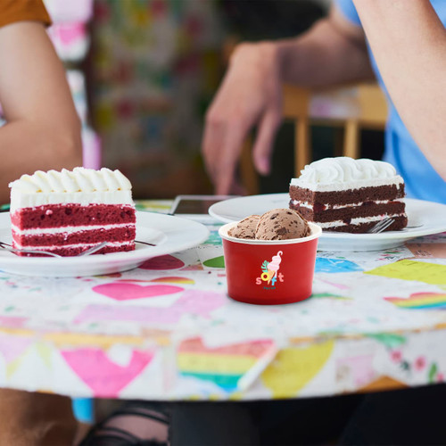 A red custom gelato cup filled with chocolate gelato sits on a table with two slices of cake.