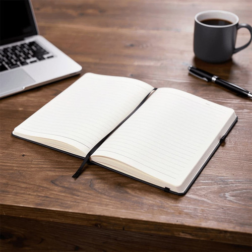 A black notebook with lined pages, open and lying on a wooden desk beside a laptop and a coffee cup.