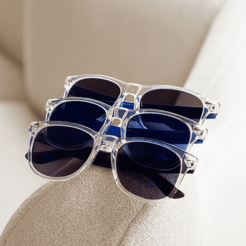 Four pairs of sunglasses sit stacked on a beige surface. The frames are clear, featuring dark blue and black lenses.