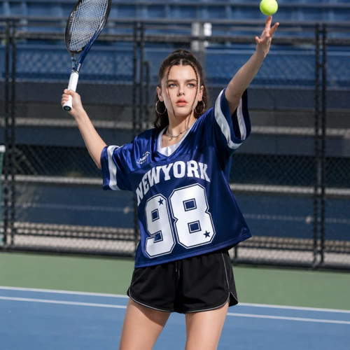 A young woman in a blue sports jersey with white text and shorts prepares to serve a tennis ball on a court.