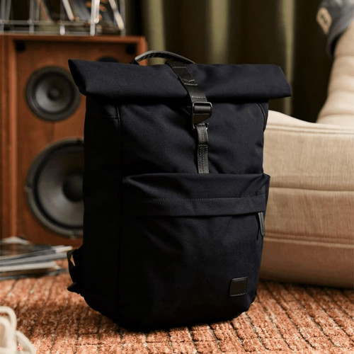 A black rolltop backpack with a front pocket and a branding logo, set against a stylish indoor backdrop.