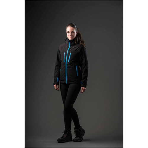 A woman wearing a black jacket with blue accents, paired with leggings and boots, standing against a dark background.