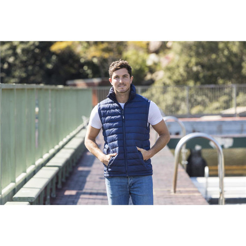 A man stands by a waterway wearing a navy blue unisex puffer vest over a white t-shirt and jeans.