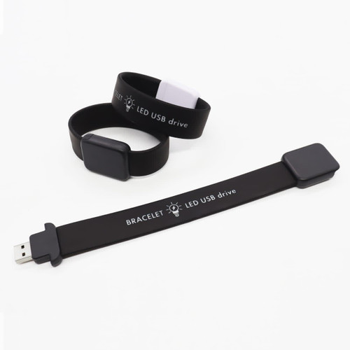 A black silicone bracelet featuring a USB drive, with an accompanying smaller black bracelet. Both have branding.