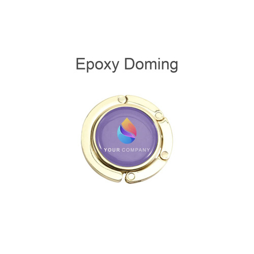 A round, foldable desk hook in gold with a purple epoxy domed centre featuring a logo.