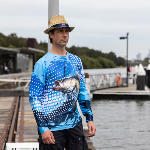 A man wearing a blue long sleeve polyester fishing tee with a fish design stands by a waterfront. He is wearing a straw hat.