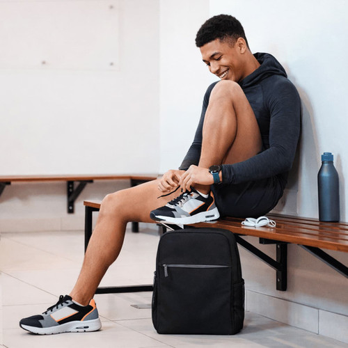 A black urban sports backpack sits on the floor beside a person tying their shoelaces in a gym setting.