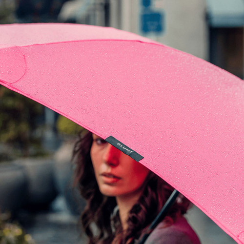 A pink umbrella with a black brand logo, partially covering a person, with raindrops on its surface.