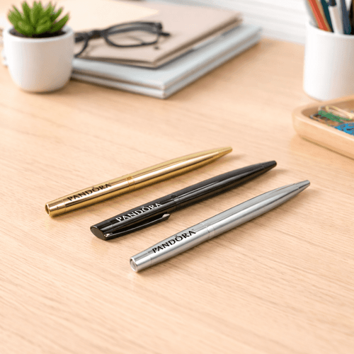 Three metal ballpoint pens in gold, black, and silver arranged on a wooden desk with stationery in the background.