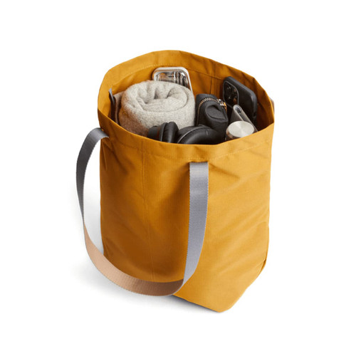 A mustard yellow tote bag filled with a blanket, headphones, and various accessories, featuring grey straps.