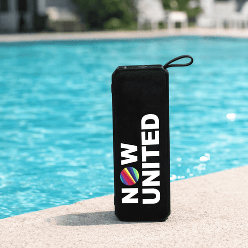 A black portable speaker with a round logo, sitting by a blue pool.