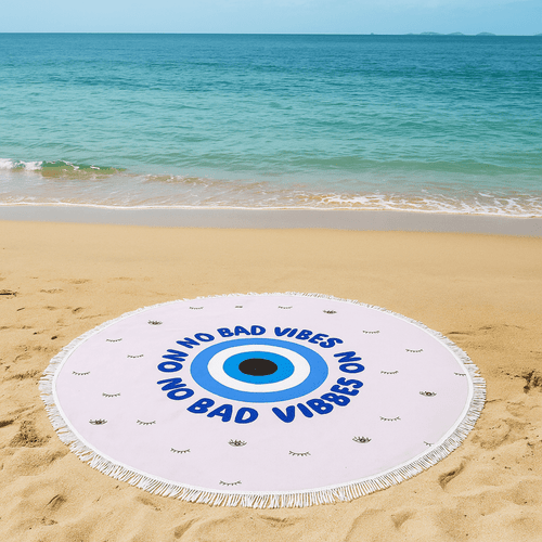 A round beach towel in white and blue with a central eye design, placed on a sandy beach by the water.