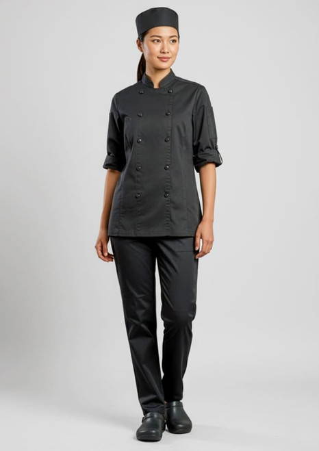 A woman in a black long sleeve chef jacket and matching pants, wearing a chef’s hat and shoes, standing against a neutral background.