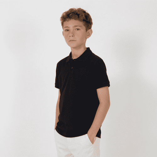 A youth black polo shirt with a collar, featuring short sleeves and a pocket. The model is standing with hands in pockets.