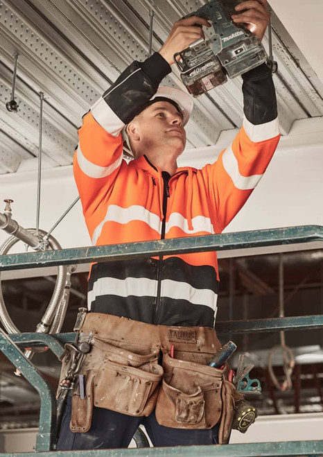 A man in an orange and black hi-vis hoodie uses a tool while standing on a scaffold, wearing a tool belt.